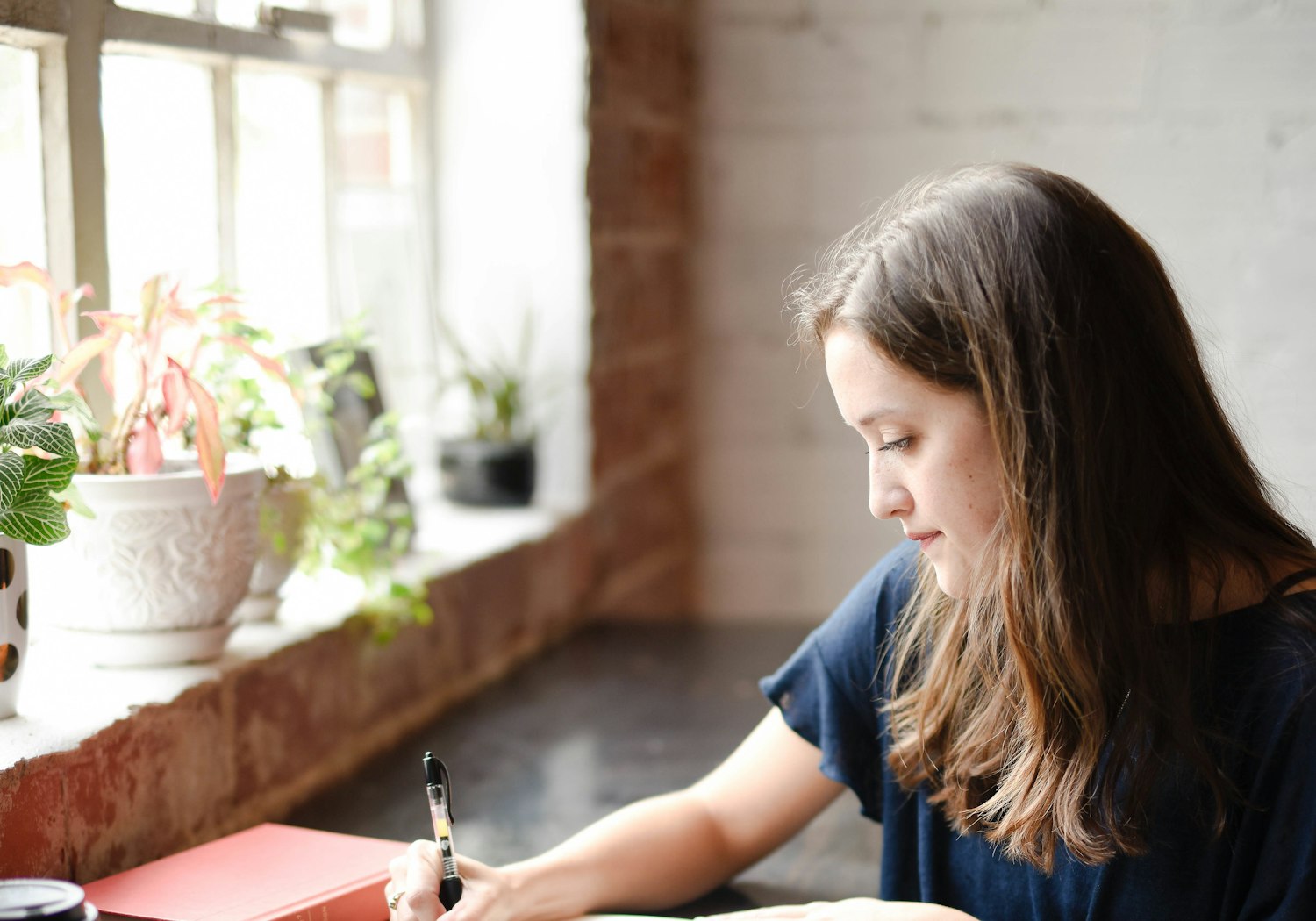 A woman writing in a notebook at a desk beside a sunlit window.