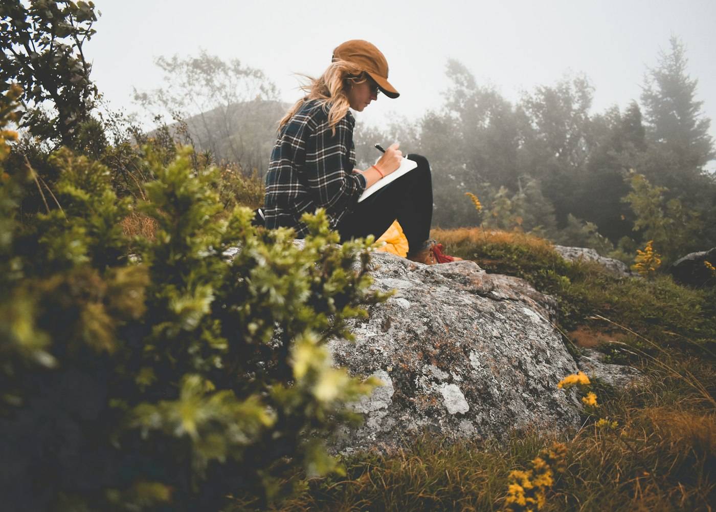 A person in a hat writing in a notebook while seated on a rock overlooking trees.