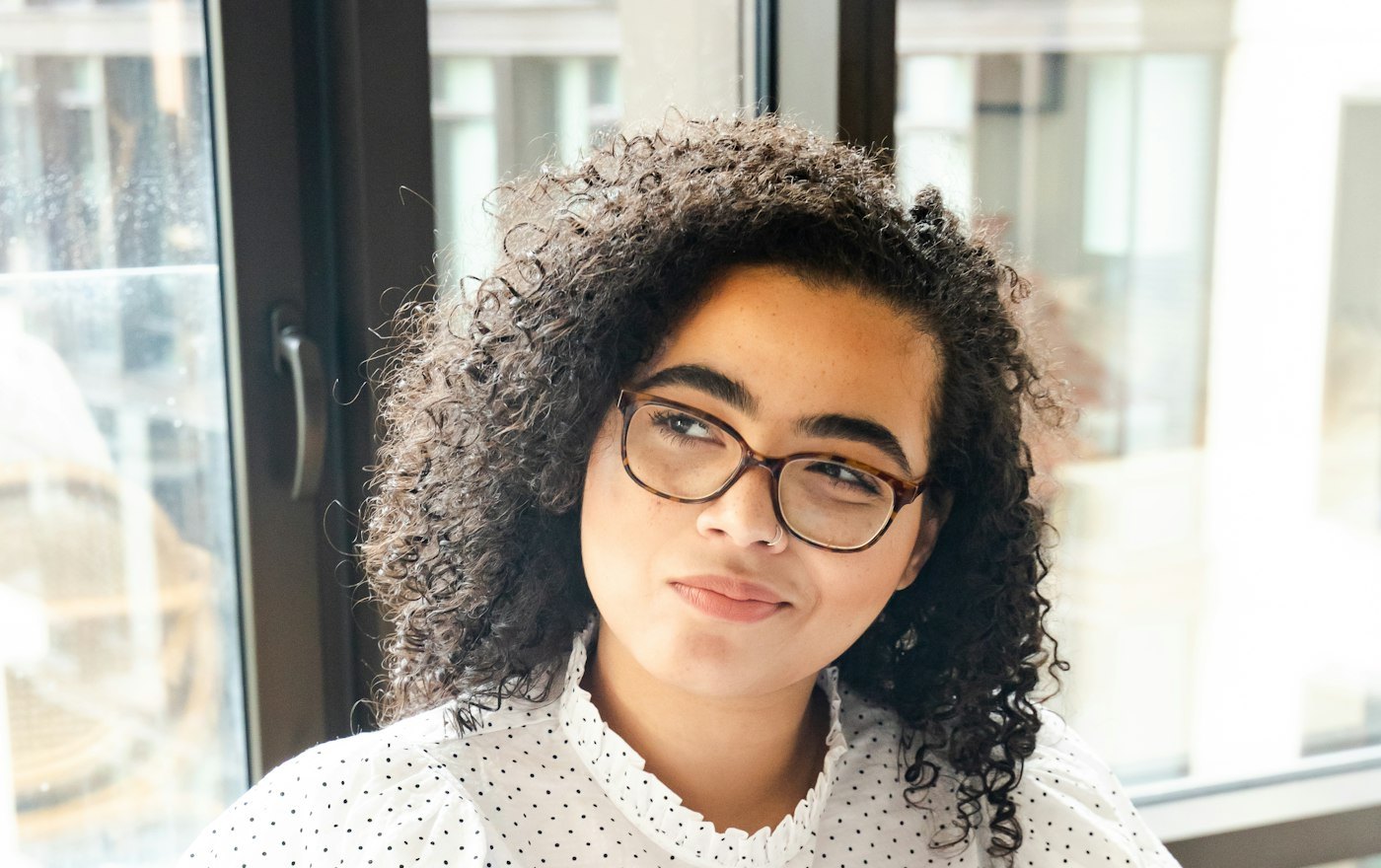 A woman with curly hair and glasses pausing thoughtfully beside a bright window.