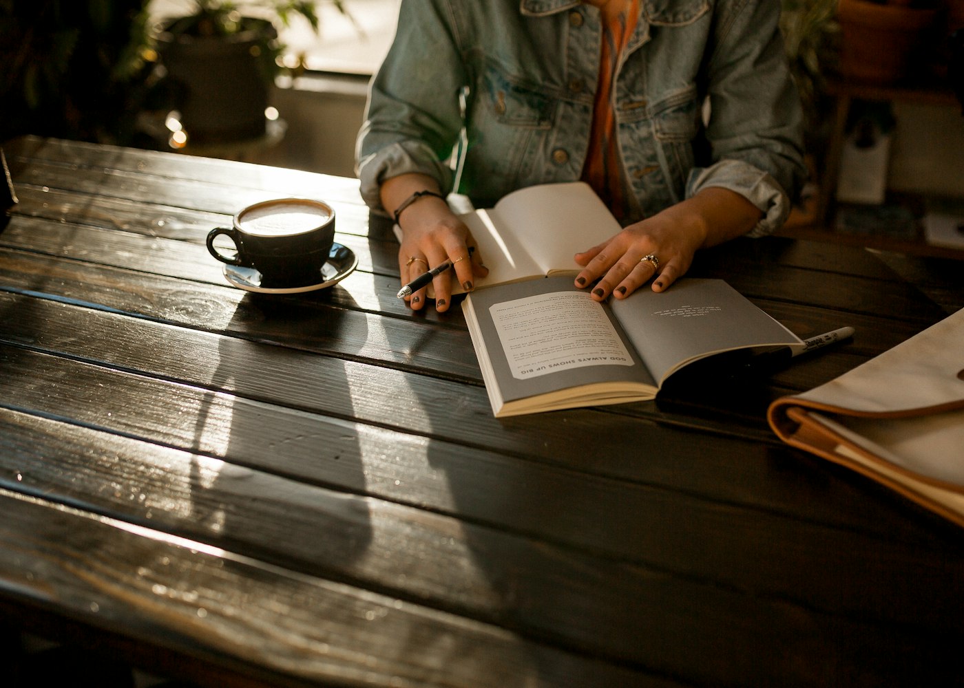 A reader with an open book, notebook and cup of coffee at a sunlit wooden table.