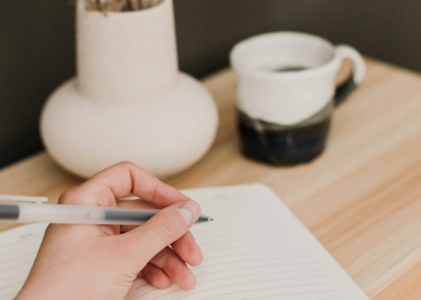 A hand holding a pen over an open lined notebook beside a cup on a wooden table.